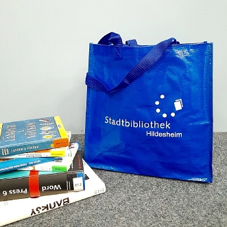 Eine gefüllte PET-Tasche in Blau mit dem Bibliothekslogo steht neben einem Bücherstapel. © Stadtbibliothek